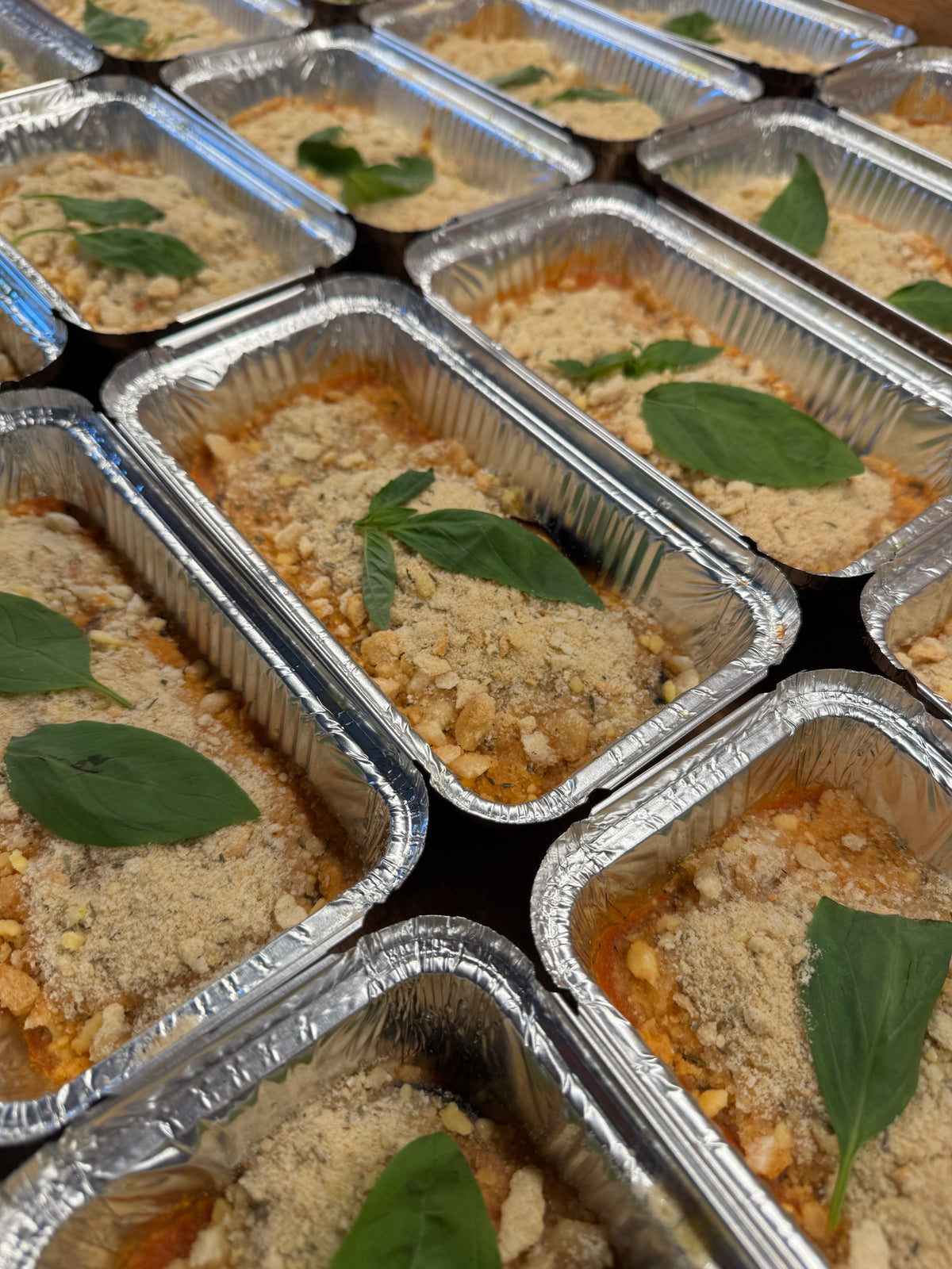 Aluminum foil containers with lasagne and topped with basil leaves.